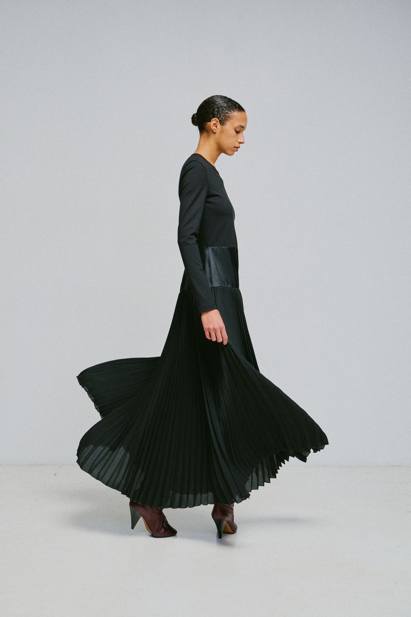 Model in a long black pleated dress spinning against a plain light grey studio background