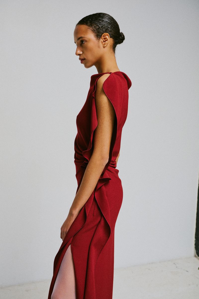 Profile view of a model in a red sleeveless dress with ruffled detailing against a white studio background.