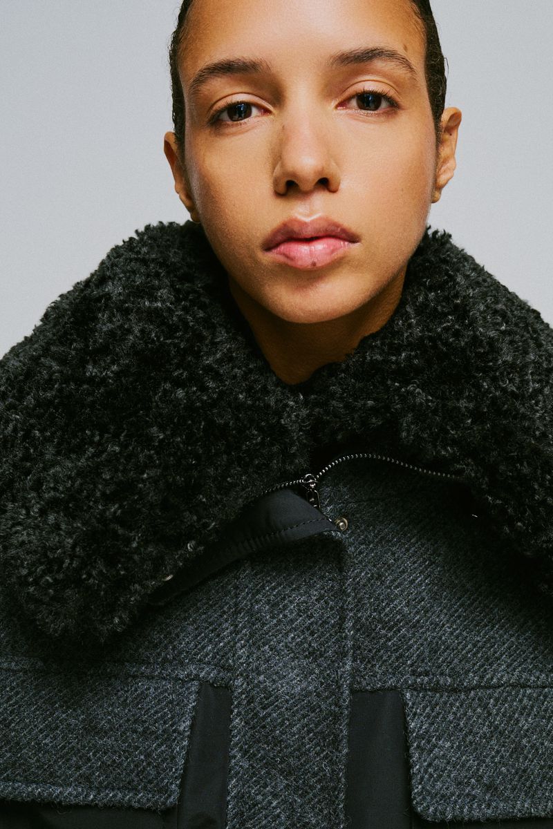 Close-up portrait of a model wearing a textured dark grey wool coat with a fluffy collar against a plain grey background
