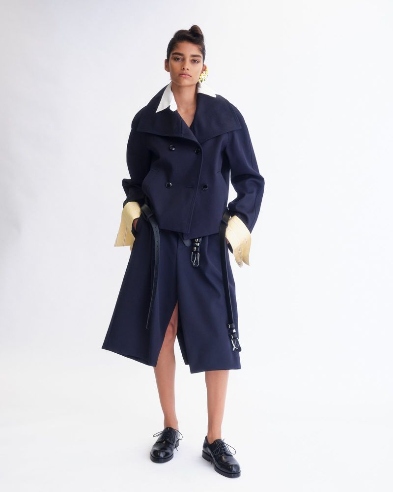 Model wearing a navy double-breasted jacket and matching midi skirt with white shirt cuffs against a white background