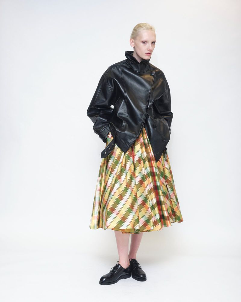 Model wearing a black leather jacket and plaid midi skirt against a plain white studio background