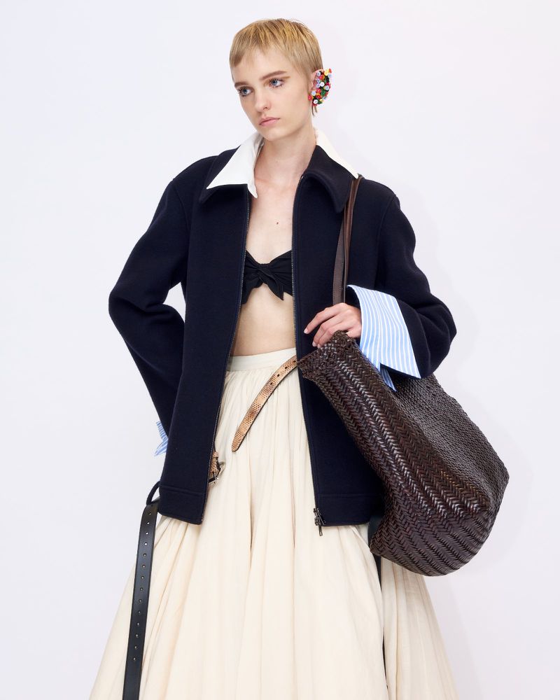 Model with short blonde hair wearing a dark zip jacket over a white collared shirt and cream skirt holding a woven brown bag against white