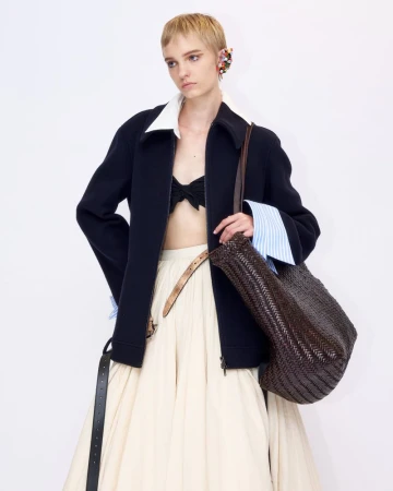 Model with short blonde hair wearing a dark zip jacket over a white collared shirt and cream skirt holding a woven brown bag against white