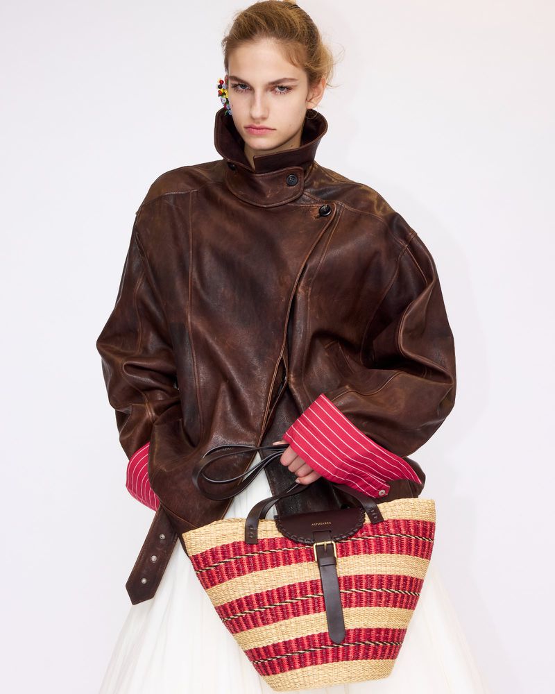 Model in dark brown leather jacket and striped pink cuffs holding a woven straw tote bag against a white background