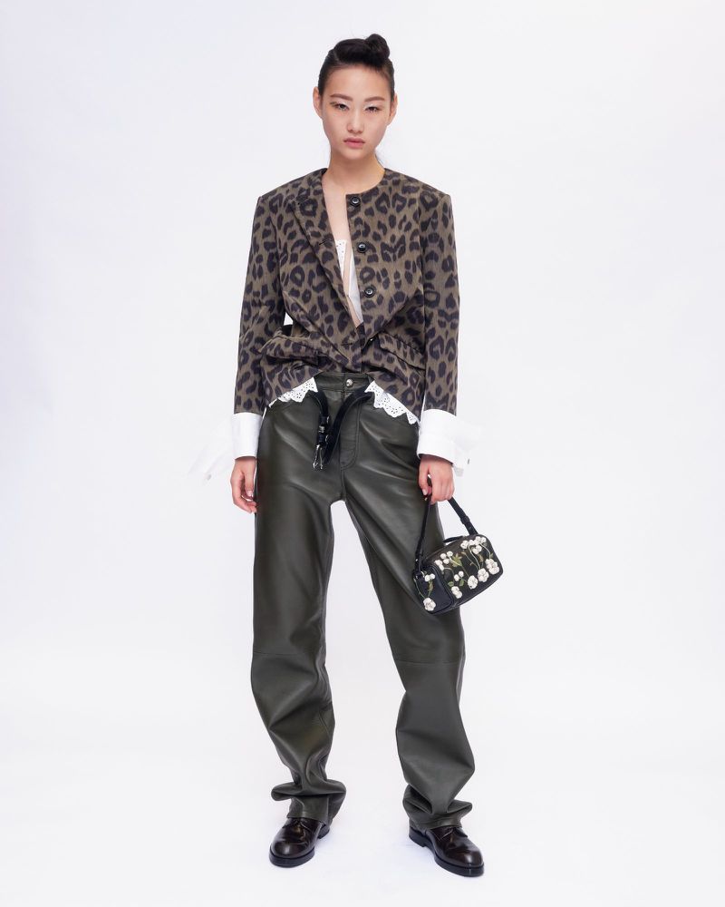Model wearing a leopard print jacket and dark leather trousers holding a small floral handbag against a white background.