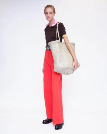 Model wearing a dark brown t-shirt and vibrant red trousers carrying a large woven tote bag against a white backdrop.