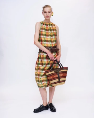 Model wearing a plaid sleeveless dress and black shoes, holding a woven handbag against a plain white background.