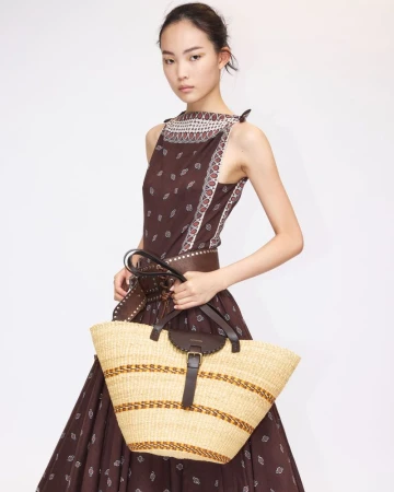 Model wearing a brown printed dress and wide studded belt holding a large straw woven tote bag against a white studio background.