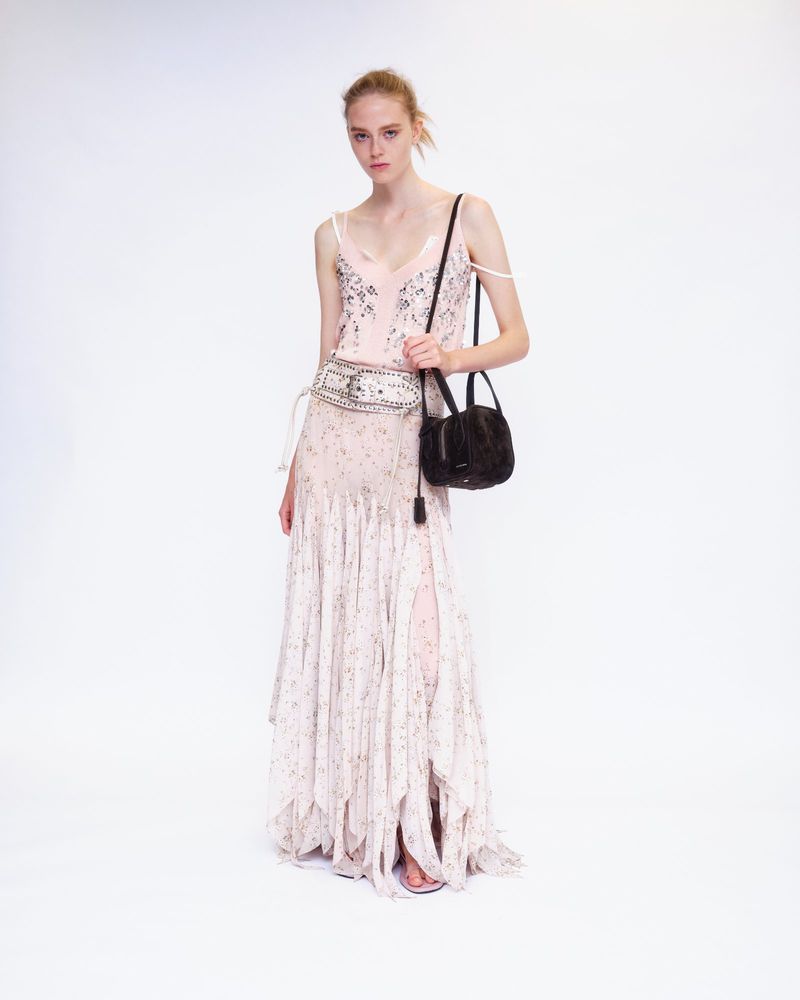 Model wearing a light pink floral dress and wide embellished belt while carrying a dark handbag against a white backdrop.