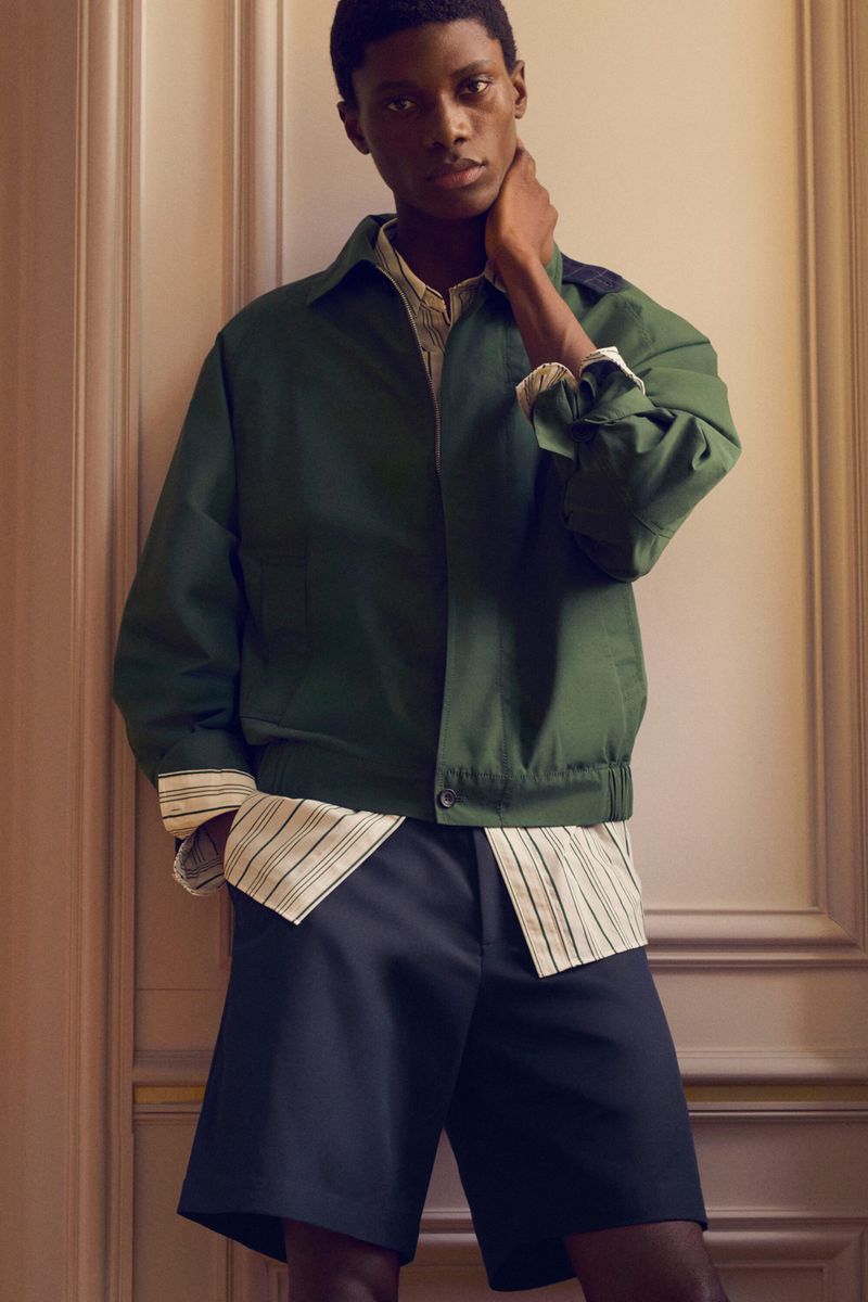 Model in green jacket and striped shirt standing against an architectural panelled wall for COS Pre-Fall 26.