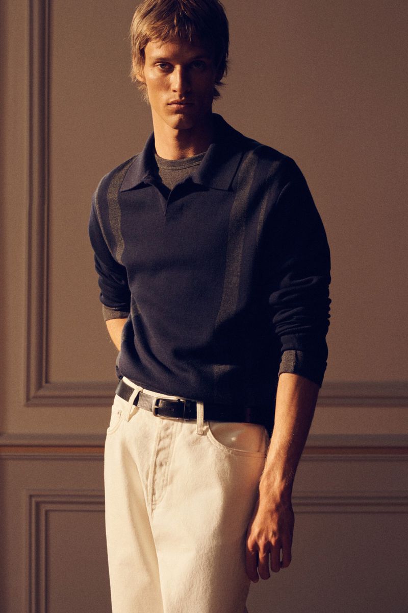 Male model in a navy polo shirt and white trousers standing against a panelled wall for COS Pre-Fall 26 lookbook