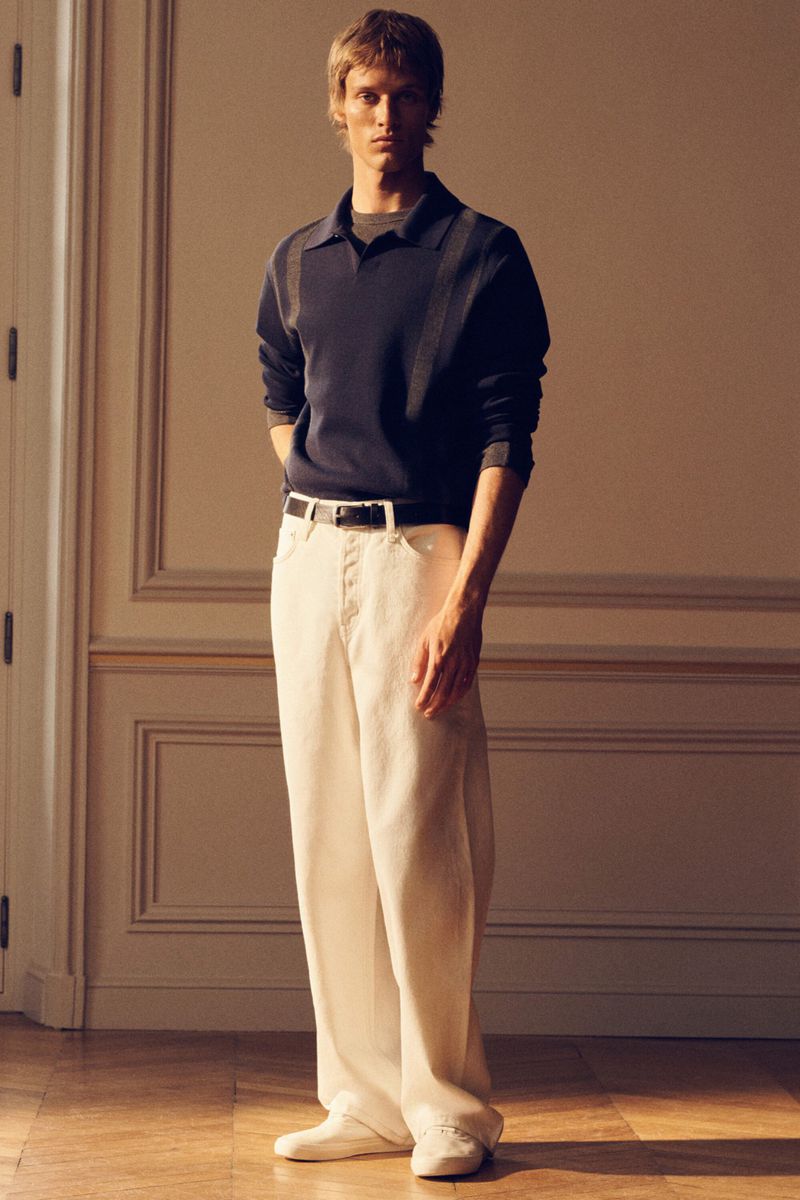 Male model in a navy polo shirt and white trousers standing against a paneled interior wall with warm lighting.