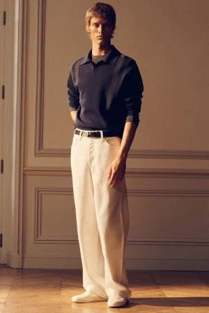 Male model in a navy polo shirt and white trousers standing against a paneled interior wall with warm lighting.