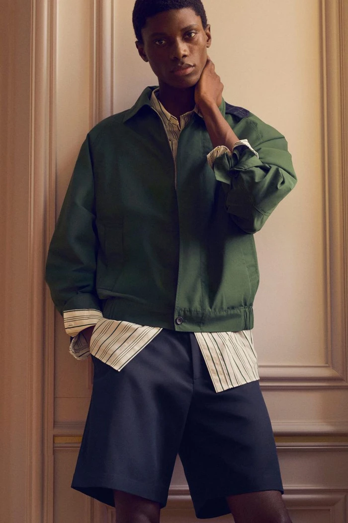 Model in green jacket and striped shirt standing against an architectural panelled wall for COS Pre-Fall 26.