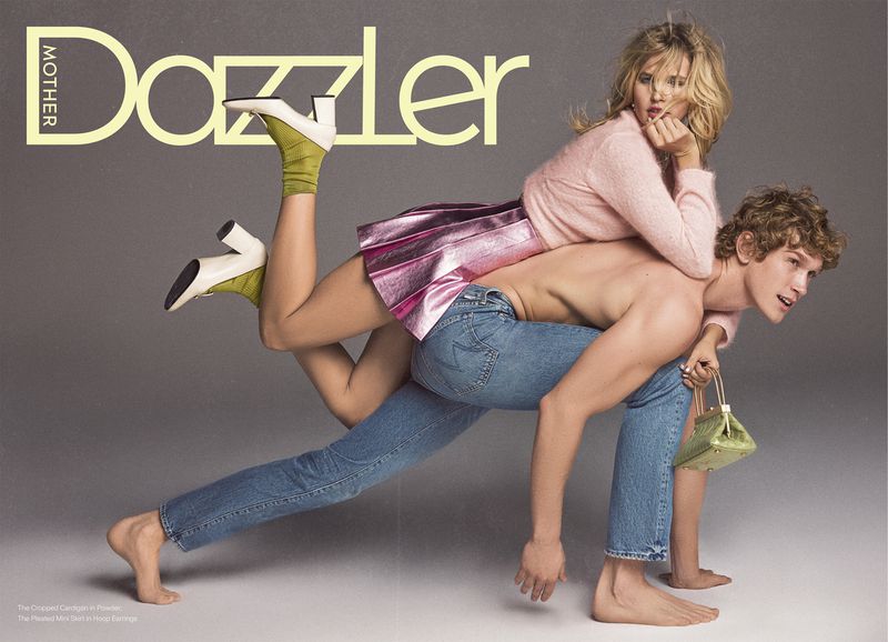 Model Jana Julius in a pink sweater and metallic skirt riding on the back of a shirtless male model wearing blue jeans in a studio setting.