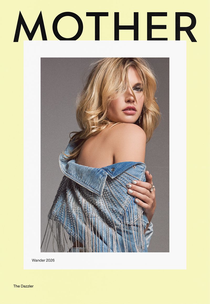 Blonde female model wearing a denim jacket with embellishments looking over her shoulder against a plain grey studio backdrop.