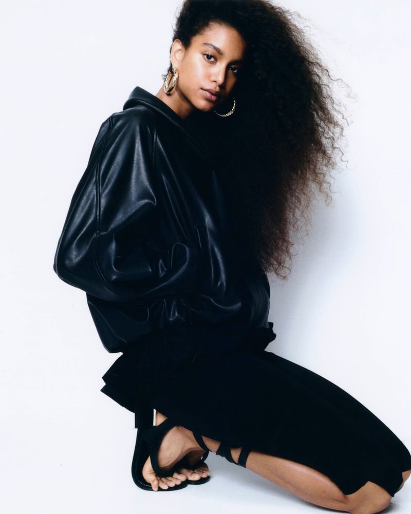 Model with voluminous dark curly hair wearing a black leather jacket and black skirt posing against white backdrop