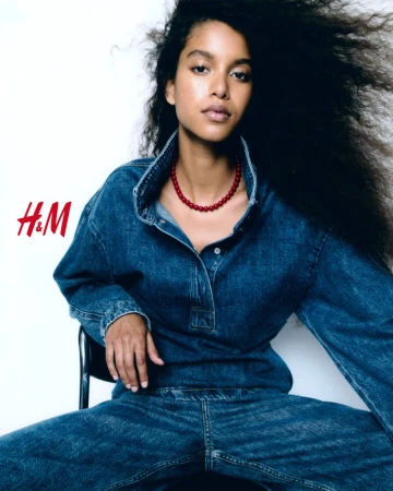 Model with long dark hair wearing a denim shirt and red bead necklace sitting against a white background