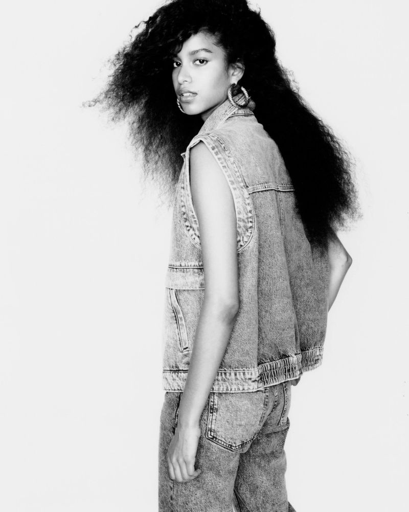 Monochrome fashion portrait of a young woman wearing a denim vest and jeans against a plain white background.