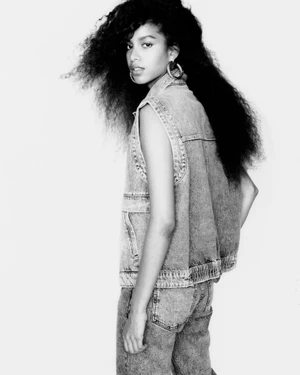 Monochrome fashion portrait of a young woman wearing a denim vest and jeans against a plain white background.