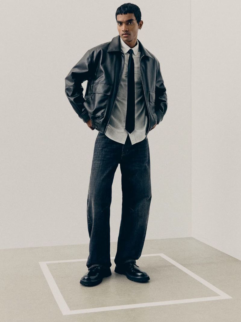 A male model wearing a black leather jacket, white striped shirt, black tie, and dark denim jeans in a white studio set.
