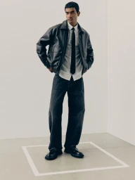 A male model wearing a black leather jacket, white striped shirt, black tie, and dark denim jeans in a white studio set.
