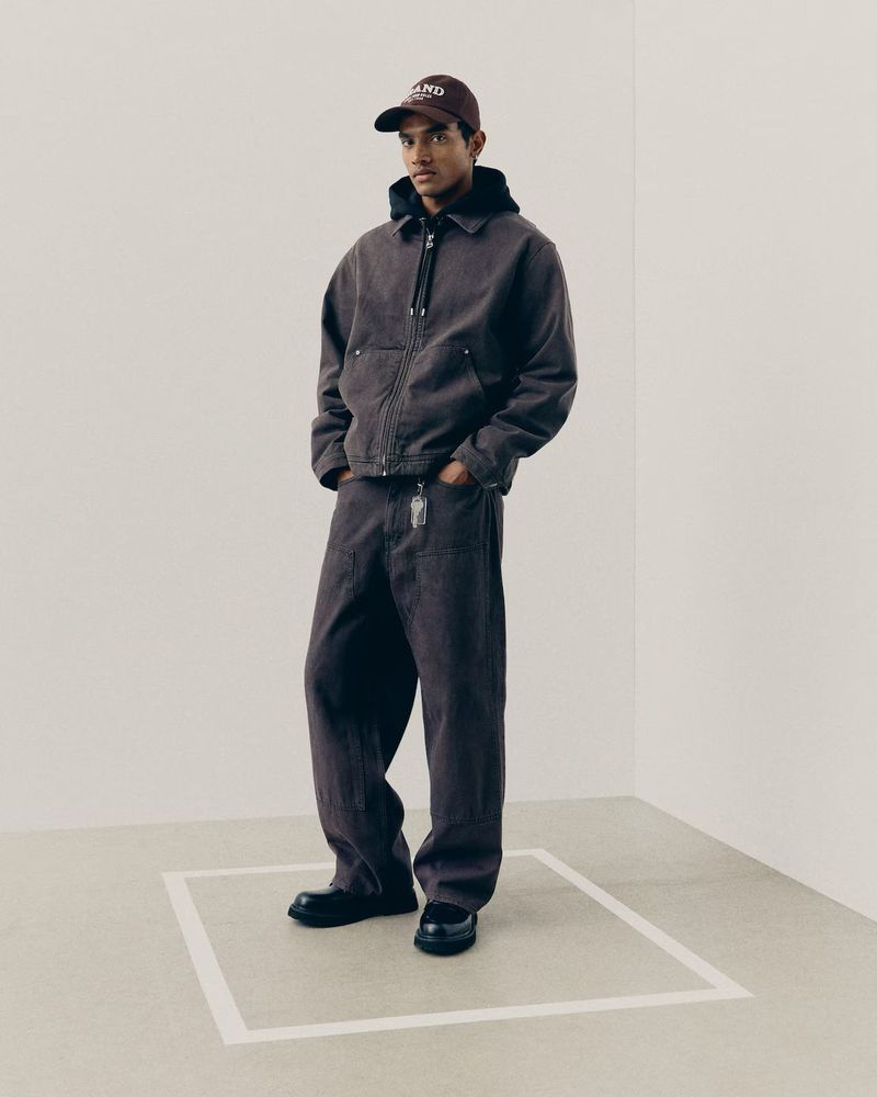 Man wearing a brown denim jacket and matching pants with a cap standing in a minimal studio setting for H&M