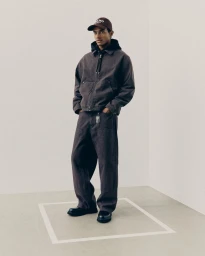 Man wearing a brown denim jacket and matching pants with a cap standing in a minimal studio setting for H&M