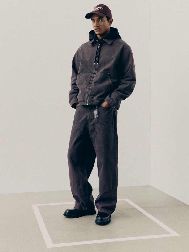 Man wearing a brown denim jacket, matching trousers, and a cap in a clean studio setting