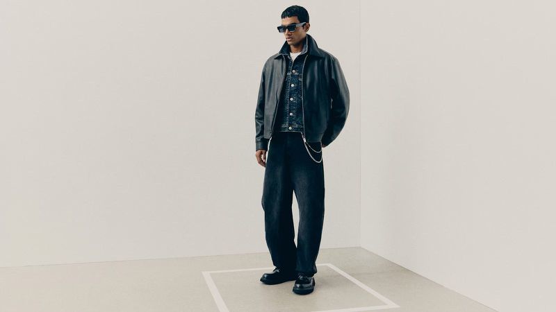 Full-length studio portrait of a male model wearing a black leather jacket, denim jacket, and dark jeans against a neutral corner backdrop