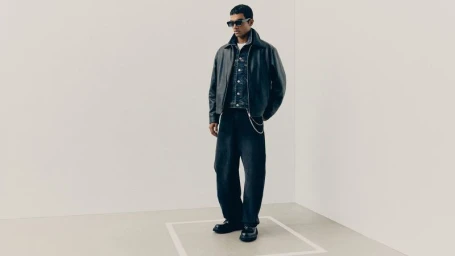 Full-length studio portrait of a male model wearing a black leather jacket, denim jacket, and dark jeans against a neutral corner backdrop