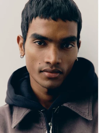 Close-up portrait of a young man wearing a dark hooded jacket against a neutral studio background.