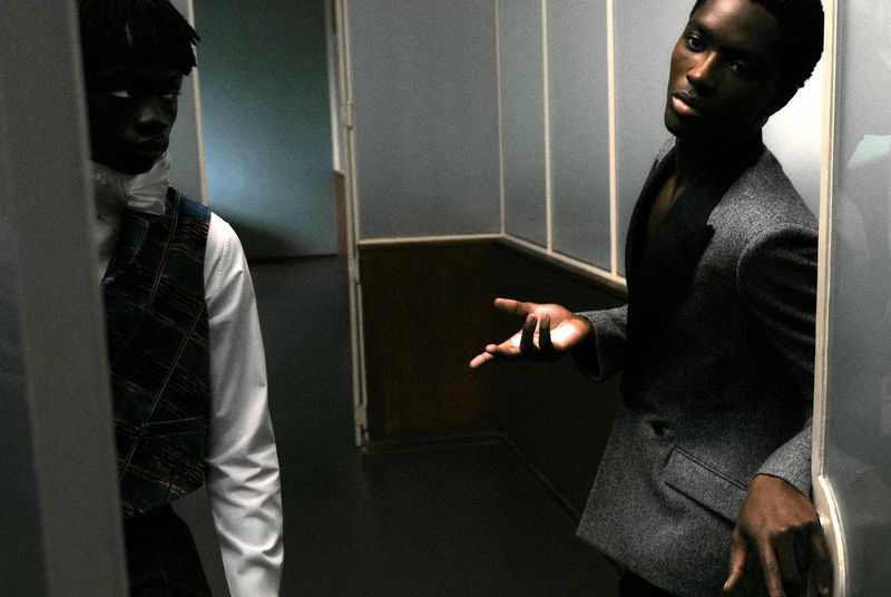Two black men in formal attire standing in a hallway, one wearing a grey blazer and gesturing with his hand.