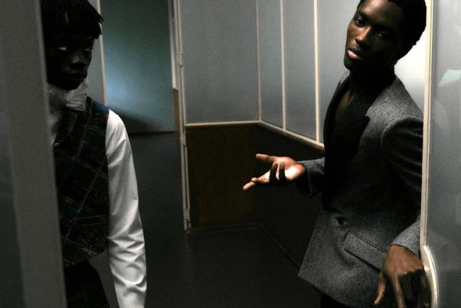 Two black men in formal attire standing in a hallway, one wearing a grey blazer and gesturing with his hand.