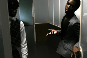 Two black men in formal attire standing in a hallway, one wearing a grey blazer and gesturing with his hand.