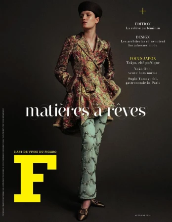 Model wearing a patterned metallic jacket and light trousers with a dark pillbox hat against a dark studio background