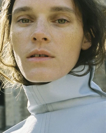 Close-up portrait of a woman with freckles wearing a light blue high collar garment outdoors in sunlight