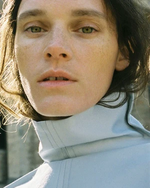 Close-up portrait of a woman with freckles wearing a light blue high collar garment outdoors in sunlight
