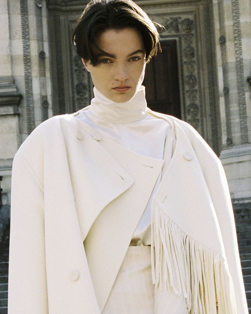 Model wearing a cream-colored high-collar coat and matching top standing in front of classical stone architecture.