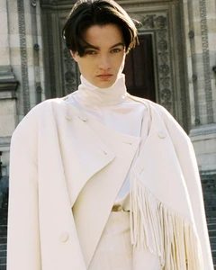 Model wearing a cream-colored high-collar coat and matching top standing in front of classical stone architecture.