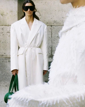 Model in white belted coat and black sunglasses holding a green handbag against a stone building exterior