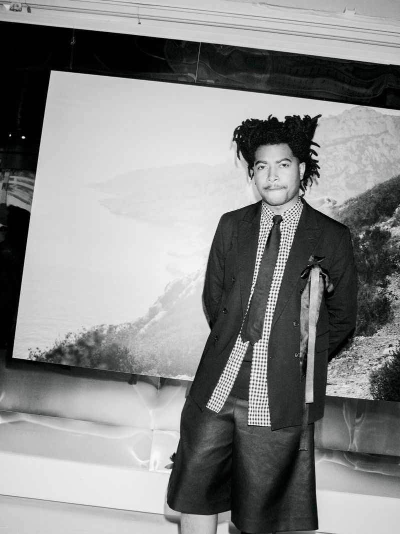 An adult male poses in a dark suit jacket and short leather pants, against a monochrome mountain background in a studio setting.