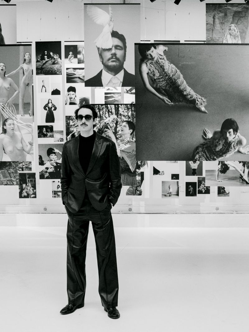 A man in a black suit stands in front of a wall of black and white photos at an art gallery, looking directly at the camera.