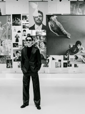 A man in a black suit stands in front of a wall of black and white photos at an art gallery, looking directly at the camera.