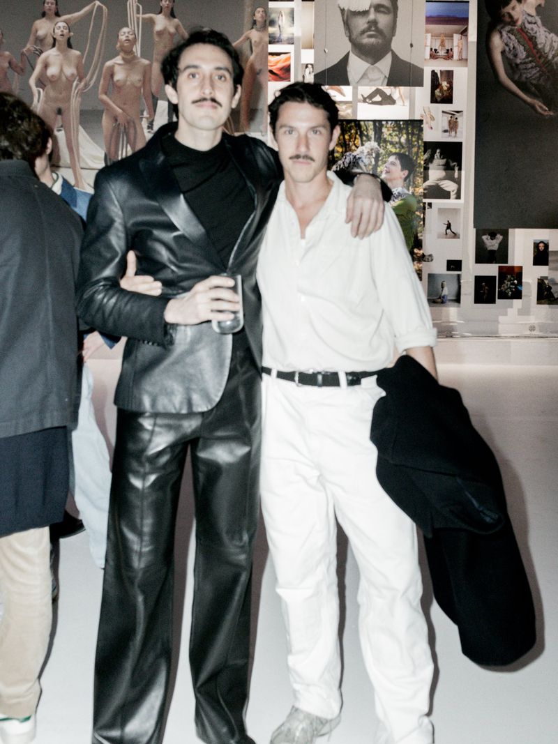Two adult men with moustaches pose indoors at an event, one in a leather suit, the other in white, standing in front of a backdrop.