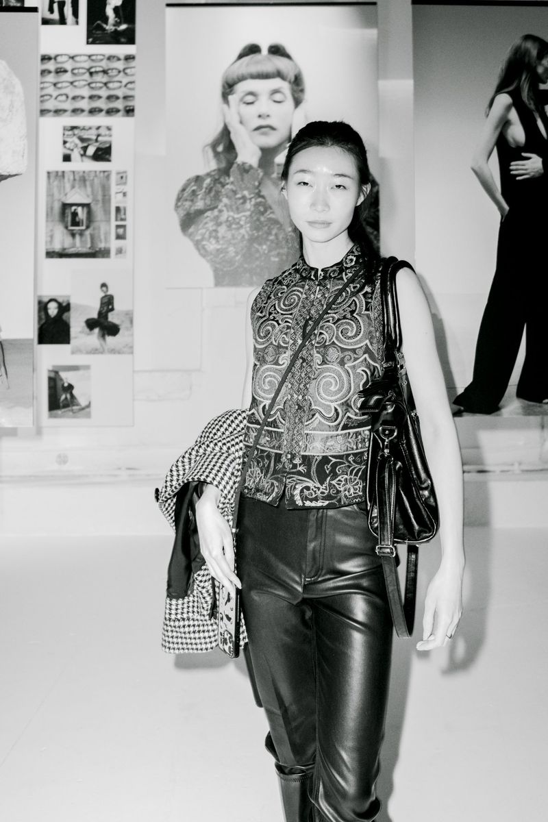 Woman in patterned vest and leather pants with a shoulder bag and jacket, posing indoors with art posters and another person visible.