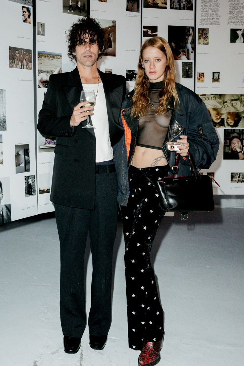 A stylish man and woman pose at an event with photo panels in the background. The woman wears a sheer top and star-patterned pants.