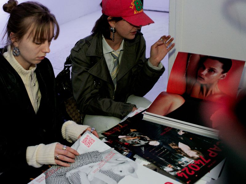 Two women seated indoors examining fashion layouts with photos, wearing contemporary clothing and accessories in a casual environment.