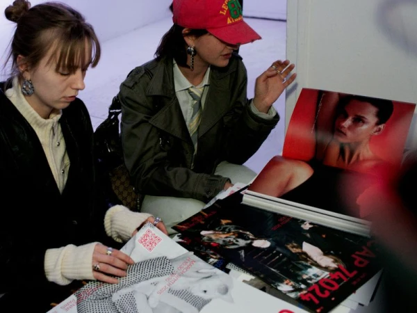 Two women seated indoors examining fashion layouts with photos, wearing contemporary clothing and accessories in a casual environment.