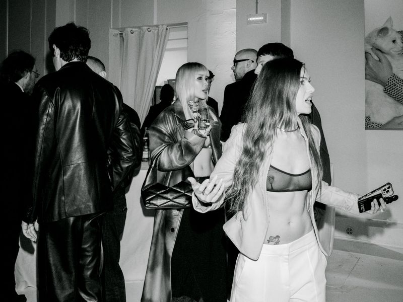 A group of people mingle indoors at an event. One woman in a white blazer holds her phone, and another holds a beverage.